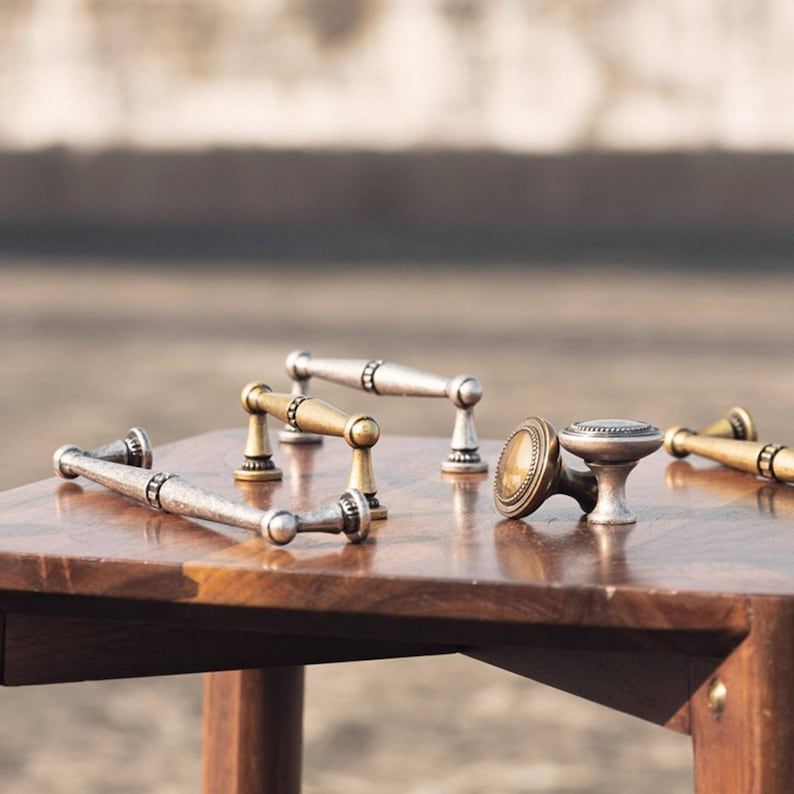 May include: A selection of cabinet handles and knobs in various metallic finishes, including silver, gold, and bronze. The hardware is displayed on a wooden surface, showcasing different designs. The image highlights the decorative nature of the hardware.