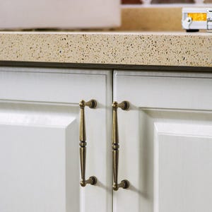May include: Two antique brass cabinet pulls with a vertical bar design are mounted on a white cabinet door. The pulls have a decorative, curved end and a detailed centre section. The countertop above is speckled beige.