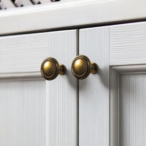 May include: A pair of antique brass cabinet knobs with a detailed, circular design. The knobs are mounted on a white cabinet door with vertical, textured lines. The knobs have a vintage, aged appearance.