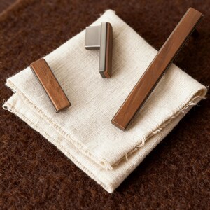 May include: A set of cabinet hardware, including a handle and two pulls, with a contemporary design. The hardware combines wood and brushed metal, displayed on a folded off-white linen cloth. The background is a textured brown surface.