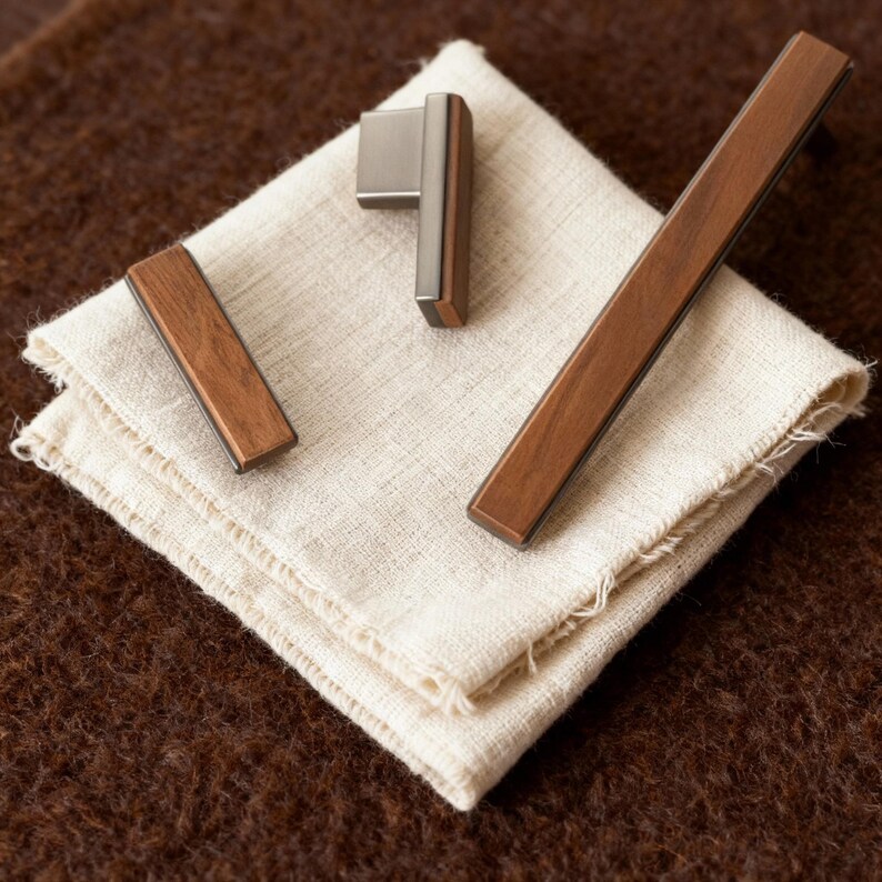 May include: A set of cabinet hardware, including a handle and two pulls, with a contemporary design. The hardware combines wood and brushed metal, displayed on a folded off-white linen cloth. The background is a textured brown surface.