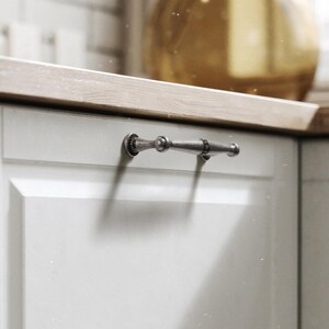 May include: A close-up of a light grey cabinet door with a silver-toned handle. The handle is a horizontal bar with decorative detailing. The countertop above is a light wood colour.