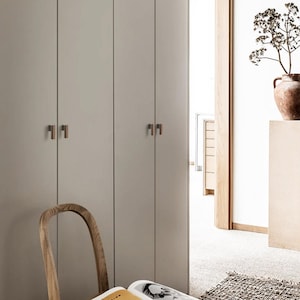 May include: A room in neutral tones featuring closed cabinets with bronze handles. A wooden chair and a vase with dried plants are also visible. The room has a minimalist design, with a focus on natural materials.