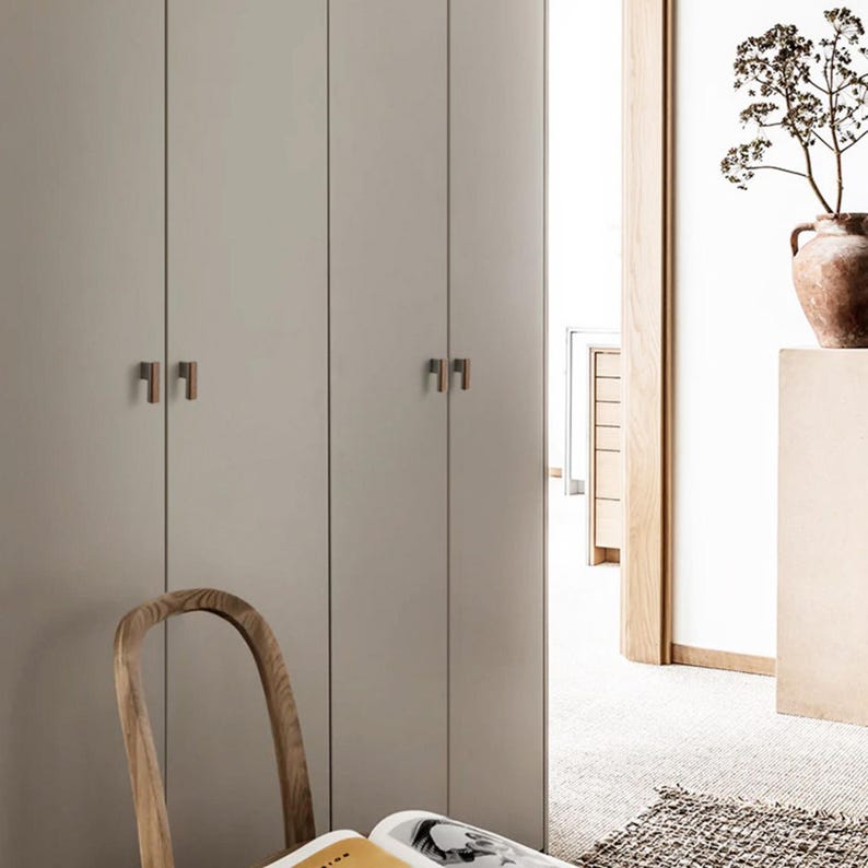 May include: A room in neutral tones featuring closed cabinets with bronze handles. A wooden chair and a vase with dried plants are also visible. The room has a minimalist design, with a focus on natural materials.