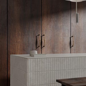 May include: A contemporary kitchen scene with dark wood cupboards and brass handles. A light grey countertop with a textured, vertical tile design is in the foreground. A small white bowl and a book are on the countertop.