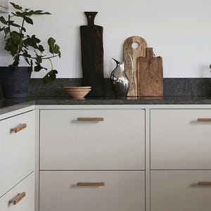 May include: A kitchen interior with a fig plant in a dark pot, wooden chopping boards, and a silver bird ornament. Light grey cabinets with wooden handles are shown. A small wooden bowl is on the countertop.