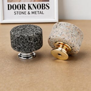 May include: Two stone door knobs with metal bases. One knob is dark gray with a speckled pattern, and the other is light gray with a speckled pattern and a gold base. The text "DOOR KNOBS STONE & METAL" is in the background.
