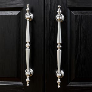 Op de afbeelding: Twee gepolijste zilveren deurgrepen met een klassiek ontwerp zijn gemonteerd op een zwarte houten deur. De handgrepen hebben een lange, slanke vorm met decoratieve details en een afgeronde boven- en onderkant.