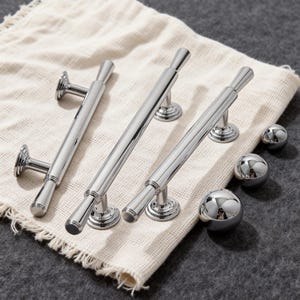May include: A collection of polished chrome cabinet pulls and spherical knobs. The pulls have a cylindrical design with flared ends and circular mounting bases. The knobs are smooth and reflective, with a mirrored surface. The hardware is displayed on a cream-colored cloth.