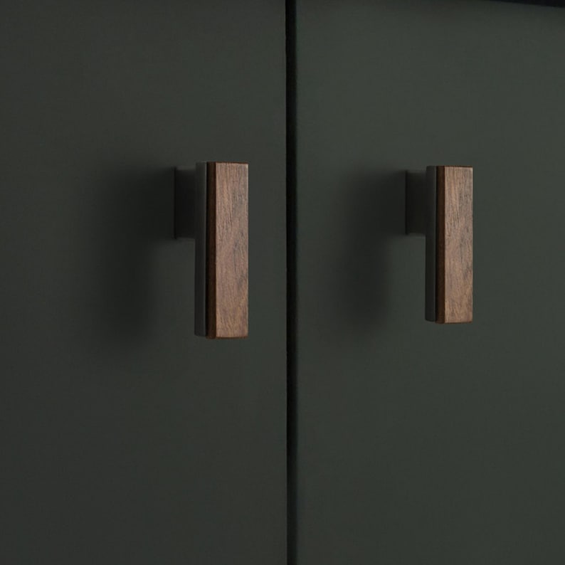 May include: Two wooden cabinet pulls with a rectangular design. The pulls are a warm brown colour and are mounted on a dark green cabinet door. The pulls are made of wood and have a simple, modern design.