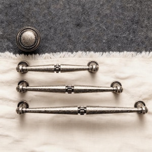 May include: A set of antique-style cabinet hardware, including a round knob and three matching pull handles. The hardware has a distressed, silver finish and is arranged on a white cloth against a gray background.