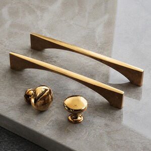 May include: Two polished gold-tone metal cabinet pulls with a curved, arching design and two matching round gold-tone cabinet knobs. Each piece features a small engraved letter M on the surface, showcasing a reflective, smooth metallic finish.