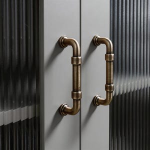 May include: Close-up of two cabinet doors with bronze-colored, pipe-shaped handles. The handles are mounted on a light gray surface, and the doors have a textured, ribbed glass panel. The hardware has a vintage, industrial aesthetic.