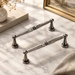 May include: Two silver-toned metal drawer pulls with a distressed finish. Each handle features a long, slender bar with a central decorative ring and two conical mounting bases. The metal surface has a textured, weathered appearance throughout.