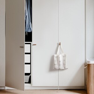 May include: A white wardrobe with a partially open door showing a blue shirt and storage. A patterned canvas tote bag hangs on the closed door. A wooden laundry basket is on the right.