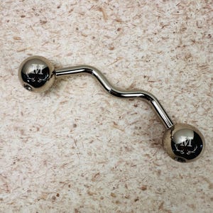 May include: A polished silver-tone metal barbell body jewelry piece with a curved shaft and two spherical ends. Each sphere is engraved with the letter M positioned above a graphic of deer antlers, creating a symmetrical design on both sides.