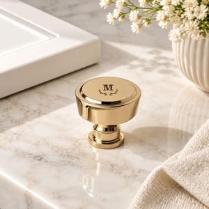 May include: A polished gold-toned metal cabinet knob with a circular top featuring an engraved letter M centred above a small antler graphic. The knob has a stepped, cylindrical base and a smooth, reflective finish, standing upright on a surface.