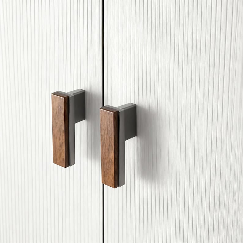 May include: Two cabinet pulls with a modern design. The pulls have a rectangular, dark wood grip attached to a dark grey metal base. They are mounted on a white cabinet door with vertical textured lines.