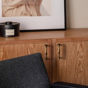 May include: A wooden cabinet with two doors and brass handles. A black round box with a label sits on top of the cabinet. A framed picture hangs above the cabinet. A dark grey upholstered chair is in the foreground.