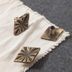Könnte beinhalten: Drei bronzefarbene Schrankknöpfe mit einem geometrischen Sternmuster. Die rautenförmigen Knöpfe haben eine strukturierte Oberfläche und einen zentralen Montagepfosten. Sie sind auf einem weißen, strukturierten Stoff angeordnet.