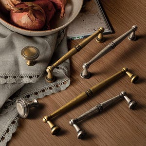 May include: A selection of antique-style cabinet hardware, featuring brass and silver-toned handles and knobs. The handles are long and slender, with decorative detailing. The knobs are round with a raised, textured design. The hardware is displayed on a wooden surface.