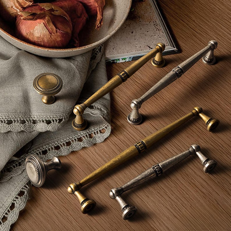 May include: A selection of antique-style cabinet hardware, featuring brass and silver-toned handles and knobs. The handles are long and slender, with decorative detailing. The knobs are round with a raised, textured design. The hardware is displayed on a wooden surface.