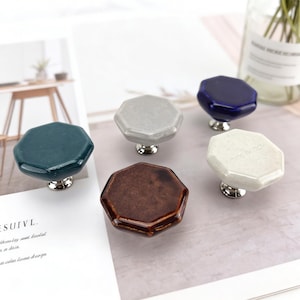 May include: Five octagonal cabinet knobs with a glossy finish in dark teal, light grey, dark blue, brown, and off-white. Each knob features a silver-toned metal base. The off-white knob has the text MADE BY TROY embossed on its top surface.