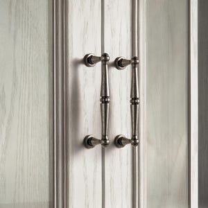 May include: A pair of antique-style cabinet door handles in a muted silver finish. The handles feature a detailed, textured design and are mounted on a white-painted wooden cabinet door with a visible wood grain.