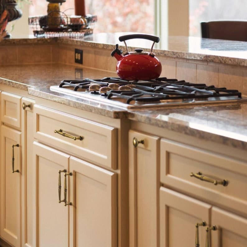 May include: A kitchen interior with a red kettle on a gas hob. The hob is set into a speckled granite worktop. Cream-coloured cabinets with brass hardware are visible below. The background includes a window and a glimpse of a dining area.