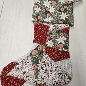 May include: A quilted Christmas stocking with a patchwork design. The stocking features various floral patterns in red, white, and green. The cuff and hanging loop have a floral print on a green background.