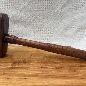 May include: A handcrafted wooden mallet with a dark brown finish. The mallet features a rectangular head and a cylindrical handle with carved grooves for grip. The tool rests on a wooden surface.