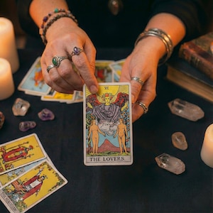 May include: A tarot card reading with the Lovers card prominently displayed. The card features a winged figure above two nude figures. Other tarot cards, crystals, and candles are arranged on a black surface, creating a mystical atmosphere.