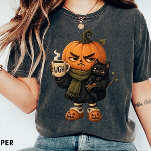 May include: A charcoal grey t-shirt featuring a graphic of a grumpy pumpkin-headed character wearing a green scarf and holding a mug with the text UGH. The character is accompanied by a small black cat and wears orange pumpkin-themed slippers.