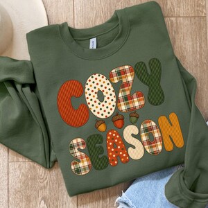 以下が含まれることがあります： オリーブグリーンのクルーネックトレーナー。COZY SEASONという文字が、オレンジのストライプ、クリーム色の水玉、赤・緑・白のチェック柄など、様々なパターンの大きな文字でデザインされています。小さな茶色のどんぐりのグラフィックがアクセントになっています。