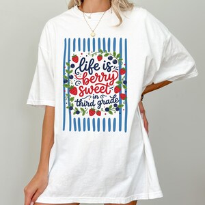 May include: A white short-sleeve t-shirt featuring a graphic print of blue vertical stripes, green vines, red strawberries, and blue berries. The text in the center reads, Life is berry sweet in third grade, written in a mix of script and print.