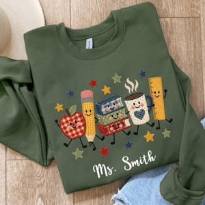May include: An olive green crewneck sweatshirt featuring an embroidered graphic of a smiling apple, pencil, stack of books, coffee mug, and ruler with arms and legs. The design includes colourful stars and the white text Ms. Smith at the bottom.