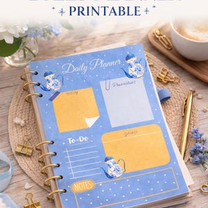 Puede incluir: Un planificador diario azul pastel con encuadernación en espiral dorada, con un diseño de tetera azul y blanca. El planificador incluye secciones para prioridades, listas de tareas y objetivos. El texto en el planificador dice "Daily Planner", "Print at Home", "Stay Organized" e "Instant Download".