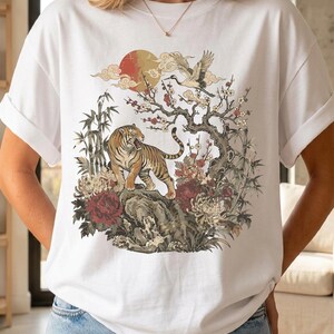 Puede incluir: Una camiseta blanca de manga corta con un estampado gráfico de un tigre sobre una roca, rodeado de bambú, flores en flor y una grulla volando. Sobre la escena, se representan un sol rojo y nubes estilizadas en una ilustración detallada.