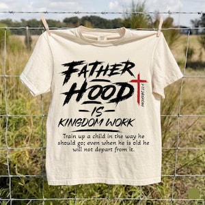 May include: A cream-colored short-sleeve t-shirt featuring black text that reads FATHER HOOD IS KINGDOM WORK with a red cross and the text PROVERBS 22:6. Below, the text reads Train up a child in the way he should go; even when he is old he will not depart from it.