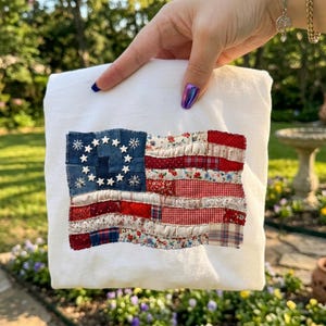 Op de afbeelding: Wit T-shirt met een patchwork Amerikaans vlagontwerp. De vlag bevat denim, bloemen en geruite stoffen in rood, wit en blauw. De vlag heeft 13 witte sterren in een cirkel op een blauwe denim achtergrond.