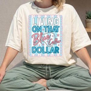 May include: Cream-colored t-shirt with the text "LIVING ON THAT Blue Collar DOLLAR" in a blue and pink design. The shirt is paired with light green pants. The overall aesthetic is casual and relaxed.