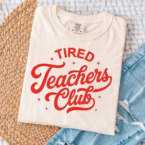 Può includere: T-shirt color crema con la scritta "Tired Teachers Club" in rosso. Il testo è circondato da piccole stelle rosse. La maglietta è piegata e posizionata su un tappetino intrecciato e denim.