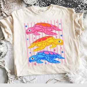 May include: Cream-colored t-shirt featuring three cartoon sea turtles in pink, yellow, and blue. Each turtle is set against a pink striped background with blue star accents. The turtles have detailed shells and friendly expressions.