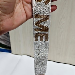 May include: White beaded strap with the words "IT'S ME" in gold beads. The strap has a gold-coloured clasp at the bottom. The strap is likely for a purse or bag.