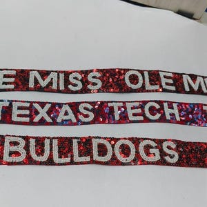 May include: Three beaded straps with gold-coloured clasps. The straps are red with white lettering. The top strap says "OLE MISS", the middle strap says "TEXAS TECH", and the bottom strap says "BULLDOGS".