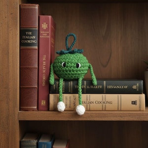 May include: A green crocheted figure with a round body, small arms and legs, and white pom-poms on the feet. The figure has black eyes and is sitting on a stack of books on a wooden shelf. The books have titles like "The Italian Cooking".