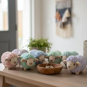 May include: A group of handmade crochet sheep in pastel shades of pink, green, blue, and lilac. The sheep are displayed on a wooden table, with a wooden bowl containing keys. A small potted plant and wall hanging are visible in the background.
