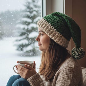 Puede incluir: Una persona con un gorro de ganchillo verde y beige con un pompón, sosteniendo una taza blanca. La persona mira por una ventana una escena nevada, con un suéter beige y vaqueros azules.