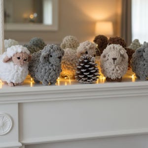 May include: A display of handmade crochet sheep in shades of white, grey, and beige, arranged on a white surface. A pine cone is placed amongst the sheep. The scene is lit by a string of small lights, creating a warm, rustic atmosphere.