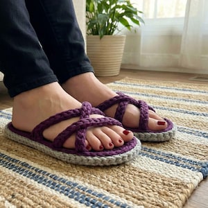 May include: Purple braided strap sandals with a grey sole. The sandals are worn on a striped rug. The sandals have a braided strap design and a comfortable, casual style. The colour scheme is a mix of purple and grey.